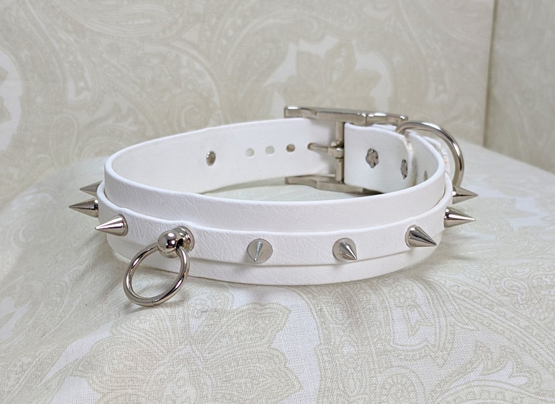 May include: White leather choker with silver spikes and a silver ring. The choker has a buckle closure.