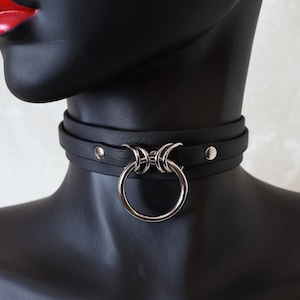 May include: A black leather choker with a silver ring and decorative accents. The choker is adjustable and has a snap closure.