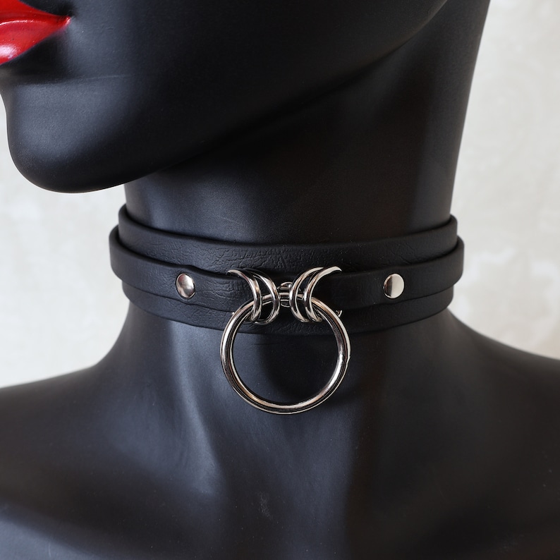 May include: A black leather choker with a silver ring and decorative accents. The choker is adjustable and has a snap closure.