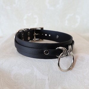 May include: A black leather dog collar with a silver ring and a silver buckle. The collar is double layered and has a textured surface.