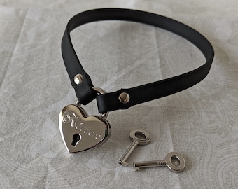 Könnte beinhalten: Ein schwarzes Lederhalsband mit einem herzförmigen Vorhängeschloss und zwei Schlüsseln. Das Vorhängeschloss trägt die Gravur "Forever".