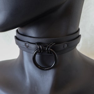 May include: A black leather choker with a large, black, circular ring. The choker has two silver-toned rivets on each side.