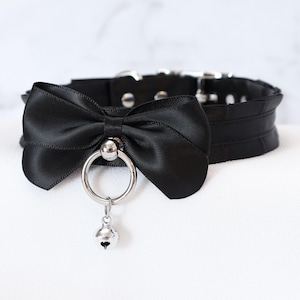 Könnte beinhalten: Schwarzes Halsband mit einer Satinschleife, einem silbernen Ring und einem kleinen Glöckchen-Anhänger. Das Halsband hat einen Schnallenverschluss und besteht aus strukturiertem Material. Die Schleife ist mittig angebracht.