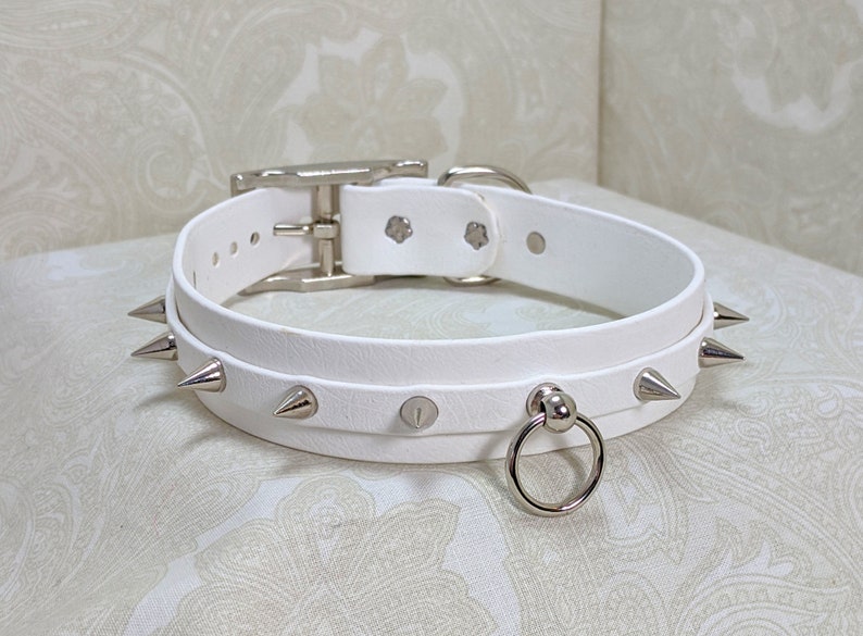 May include: White leather choker with silver spikes and a silver ring. The choker has a buckle closure.