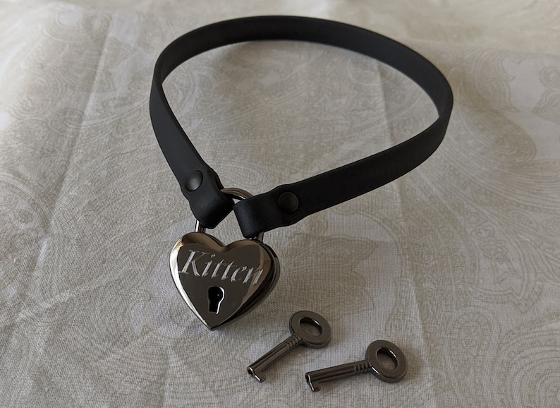 K&ouml;nnte beinhalten: Schwarzer Choker mit einem herzf&ouml;rmigen Vorh&auml;ngeschloss, auf dem "Kitten" steht, und zwei silbernen Schl&uuml;sseln.