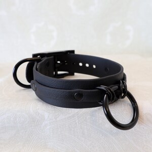 May include: Black leather choker with a metal ring and buckle closure. The choker has a textured surface and is adjustable.