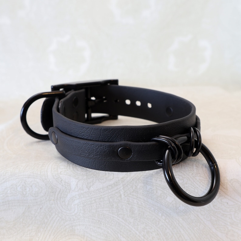 May include: Black leather choker with a metal ring and buckle closure. The choker has a textured surface and is adjustable.