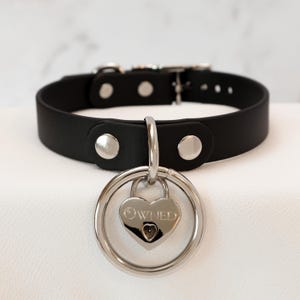 May include: Black leather collar featuring a silver heart-shaped padlock pendant. The pendant is engraved with the word "OWNED" and hangs from a silver ring. The collar has a buckle closure and silver rivets.