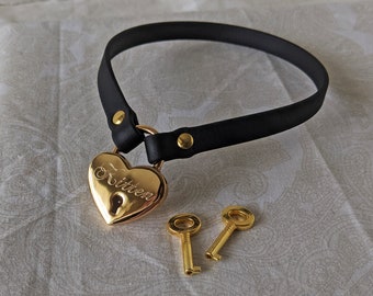 Könnte beinhalten: Schwarze Leder-Halskette mit herzförmigem goldenem Vorhängeschloss. Das Vorhängeschloss ist mit dem Wort "Kitten" graviert und enthält zwei goldene Schlüssel. Die Halskette ist mit goldfarbenen Nieten befestigt.