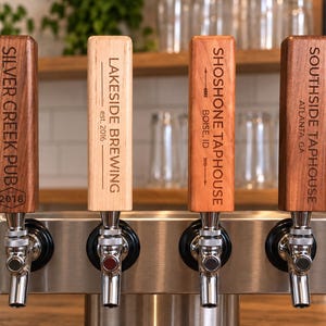 May include: Four rectangular wooden beer tap handles with metal faucets mounted on a stainless steel base. Each handle features engraved text: SILVER CREEK PUB 2018, LAKESIDE BREWING est. 2016, SHOSHONE TAPHOUSE BOISE, ID, and SOUTHSIDE TAPHOUSE ATLANTA, GA.