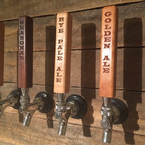 May include: Four wooden beer tap handles with different beer names engraved on them. The handles are mounted on a wooden wall. The names are "Child's Brew", "Seasonal", "Rye Pale Ale", and "Golden Ale".