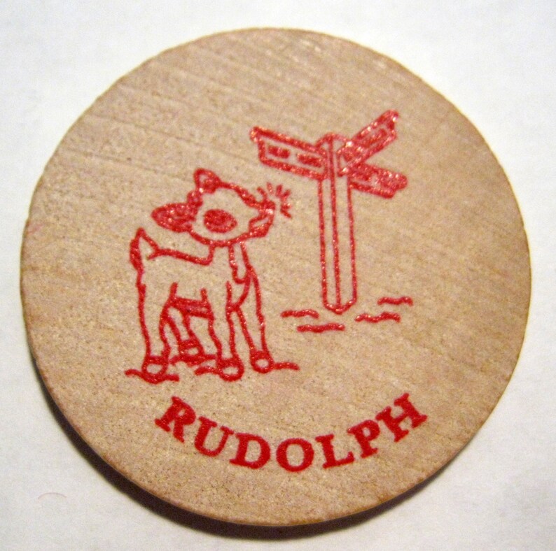 Puede incluir: Una ficha de madera roja con una imagen de dibujos animados de un reno de pie cerca de un poste indicador rojo. El texto "RUDOLPH" est&aacute; impreso en rojo debajo de la imagen.