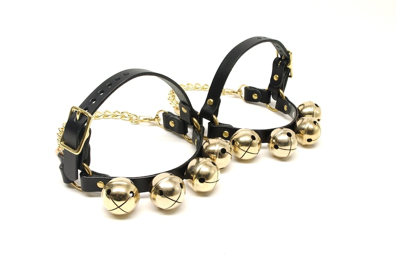 May include: Two black leather collars with gold-colored bells and hardware. Each collar has a buckle closure and is connected by a gold chain. The bells are round and have a shiny finish.
