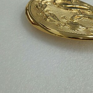 K&ouml;nnte beinhalten: Eine goldfarbene G&uuml;rtelschnalle aus Metall mit einem Anker-Design, das von einem Lorbeerkranz umgeben ist.
