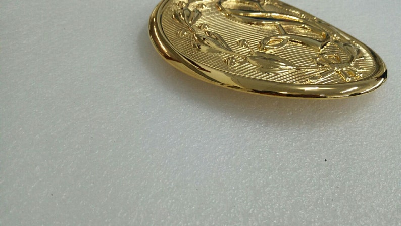 K&ouml;nnte beinhalten: Eine goldfarbene G&uuml;rtelschnalle aus Metall mit einem Anker-Design, das von einem Lorbeerkranz umgeben ist.