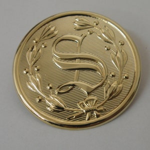 May include: A gold-toned button featuring a raised, stylised letter "S" encircled by a wreath of leaves and berries.