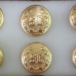 May include: Six gold-colored buttons with the letters "SC" in the center, surrounded by a laurel wreath design. The buttons are round and have a shiny finish, presented in a white box.
