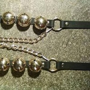 May include: Two black leather anklets with gold-colored bells and metal hardware. Each anklet has a buckle closure and a chain connecting to the bells. The bells are evenly spaced along the length of the anklets.
