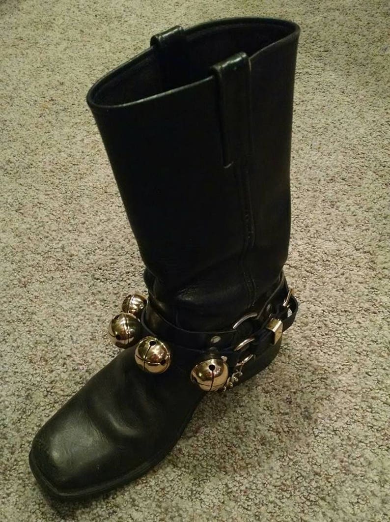 May include: A black leather boot with a square toe and a decorative strap around the ankle. The strap features several gold-colored bells and silver-colored buckles. The boot is set against a neutral carpet.