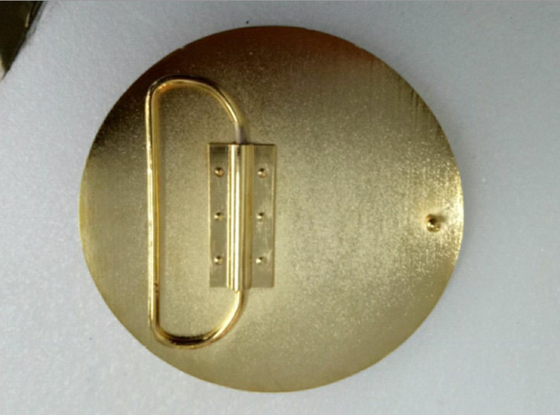 K&ouml;nnte beinhalten: Goldfarbener G&uuml;rtelverschluss aus Metall mit runder Form und einem Scharnierverschluss. Die Schnalle hat eine glatte, leicht strukturierte Oberfl&auml;che.