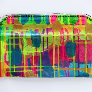 May include: A colorful clutch purse with a clear plastic front and a black stitched border. The front of the purse features a geometric pattern of bright colors, including yellow, green, pink, and blue, with gold accents.