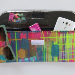 May include: A clear plastic clutch bag with a colorful abstract design. The bag has a black flap with a white phone and a pink package inside. A pair of green sunglasses are on the outside of the bag.