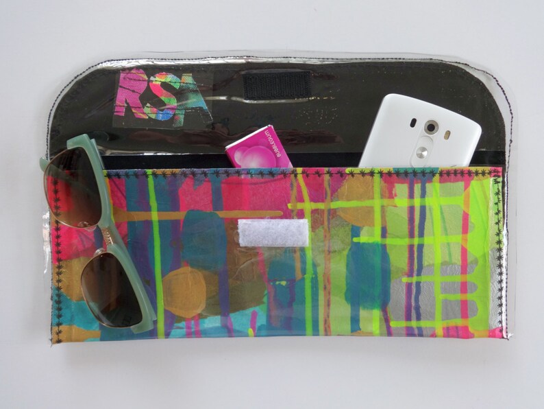 May include: A clear plastic clutch bag with a colorful abstract design. The bag has a black flap with a white phone and a pink package inside. A pair of green sunglasses are on the outside of the bag.