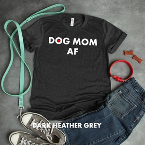 May include: Dark heather grey t-shirt with the text "DOG MOM AF" in white lettering. The shirt is shown with a red dog collar, a blue jean, a pair of sneakers, and a teal leash.