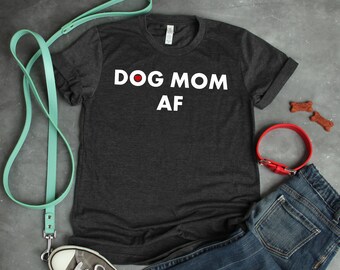 May include: Dark heather grey t-shirt with the text "DOG MOM AF" in white lettering. The shirt is shown with a red dog collar, a blue jean, a pair of sneakers, and a teal leash.