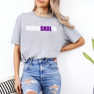 Op de afbeelding: Een grijs T-shirt met de tekst "MINNESKOLTA" in witte letters met een paarse omtrek.