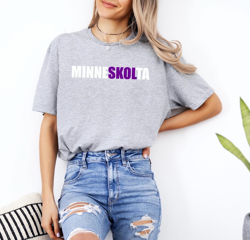 Op de afbeelding: Een grijs T-shirt met de tekst "MINNESKOLTA" in witte letters met een paarse omtrek.