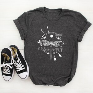 May include: A dark gray t-shirt with a white graphic design of a dragonfly with a circle around it. The circle has moons and arrows around it. The text "DRAGONFLY" is below the dragonfly.