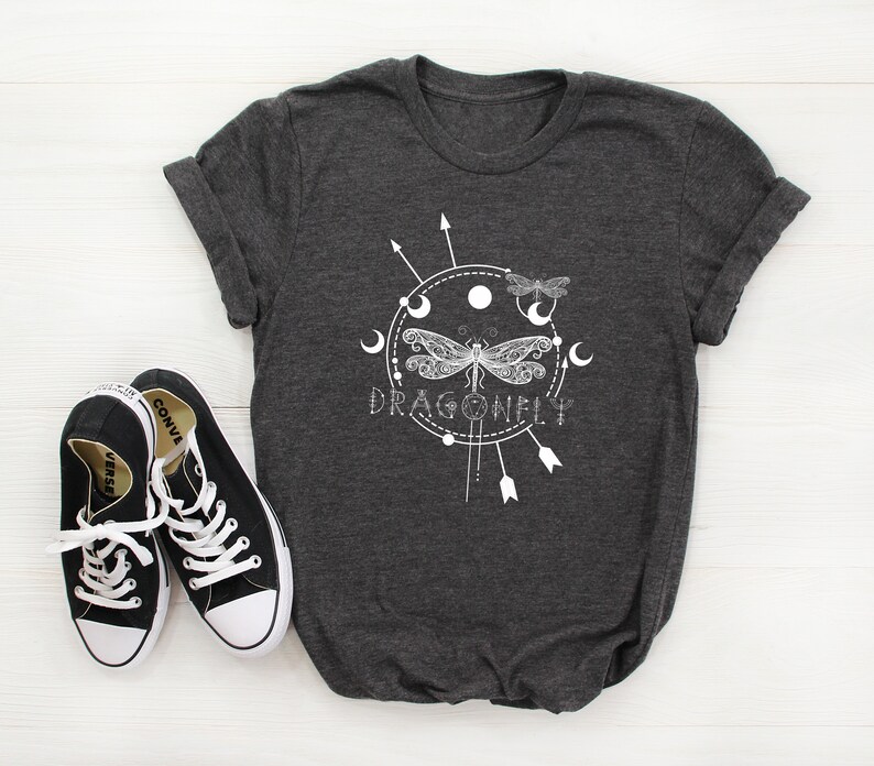 May include: A dark gray t-shirt with a white graphic design of a dragonfly with a circle around it. The circle has moons and arrows around it. The text "DRAGONFLY" is below the dragonfly.