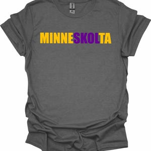 Op de afbeelding: Een donkergrijs T-shirt met de tekst "MINNESKOLTA" in gele en paarse letters.