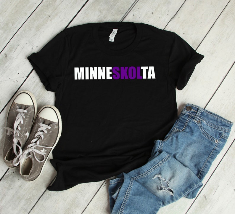 Op de afbeelding: Zwart T-shirt met witte tekst die "MINNESKOLTA" in paarse letters leest. Het shirt ligt plat op een wit houten oppervlak met een paar grijze sneakers en een blauwe jeans.