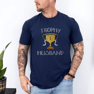 May include: A navy blue t-shirt with a distressed white graphic of a gold trophy with the number "1" on it and the text "Trophy Husband".