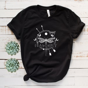 May include: Black t-shirt with a white graphic design of a dragonfly with intricate details, surrounded by moons and arrows. The text "DRAGONFLY" is printed below the dragonfly.