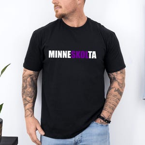 Op de afbeelding: Een zwart T-shirt met de tekst "MINNESKOLTA" in wit gedrukt, waarbij het gedeelte "KOLTA" in paars is.