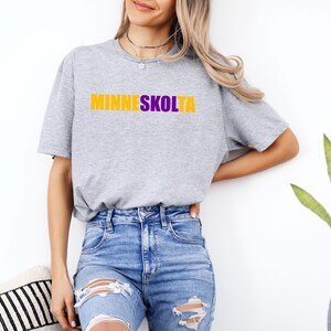 Op de afbeelding: Een grijs T-shirt met de tekst "MINNESKOLTA" in gele en paarse letters.