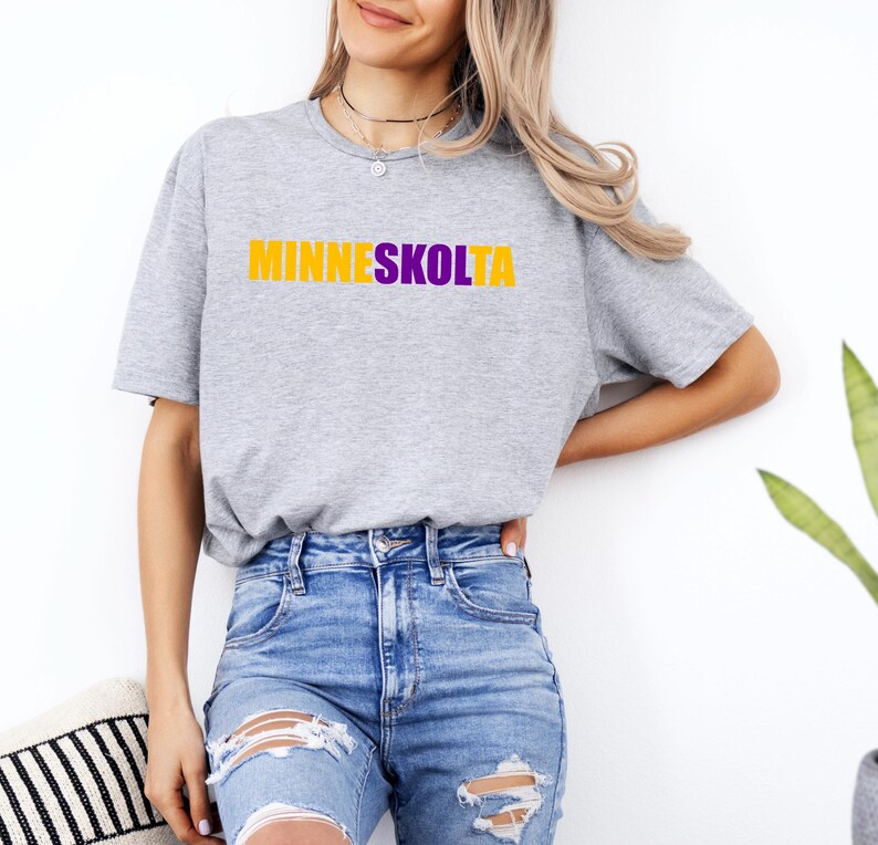 Op de afbeelding: Een grijs T-shirt met de tekst "MINNESKOLTA" in gele en paarse letters.