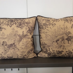 Op de afbeelding: Twee decoratieve kussens met een bruin en beige toile-patroon met jachthonden, fazanten en bladeren. De kussens hebben een bruine rand.