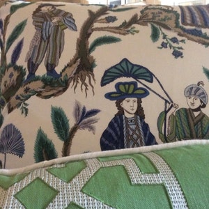 May include: Two decorative pillows with a white background and a green, blue, and brown floral pattern. The pillow on top features a stylized image of two people wearing hats and holding a large leaf. The bottom pillow is a solid green color with a white geometric pattern.