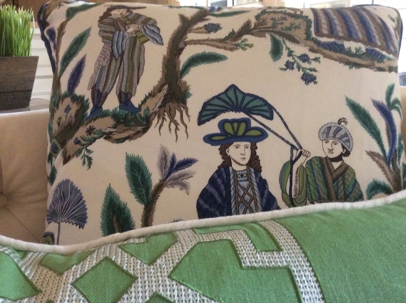 May include: Two decorative pillows with a white background and a green, blue, and brown floral pattern. The pillow on top features a stylized image of two people wearing hats and holding a large leaf. The bottom pillow is a solid green color with a white geometric pattern.
