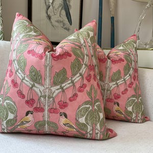 May include: Two pink throw pillows with a floral pattern featuring cherry blossoms and birds. The pillows have a white background with pink cherry blossoms and green leaves. The birds are a light brown color with yellow accents.