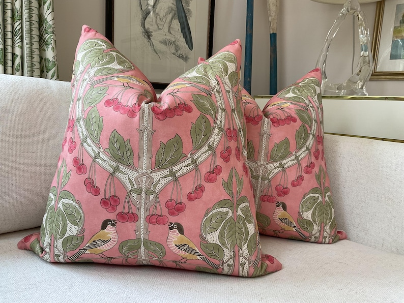 May include: Two pink throw pillows with a floral pattern featuring cherry blossoms and birds. The pillows have a white background with pink cherry blossoms and green leaves. The birds are a light brown color with yellow accents.
