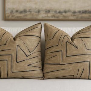 May include: Two square throw pillows featuring a tan base color with a hand-painted style black geometric line pattern. The design consists of angular, interconnected lines that create a series of abstract shapes across the fabric surface.