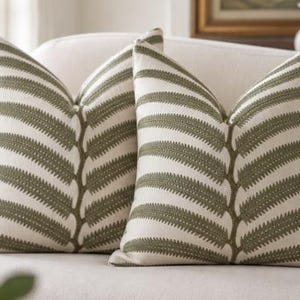 May include: Two square throw pillows featuring an off-white base fabric with a centered, vertical olive green embroidered fern leaf pattern. The embroidery extends horizontally across the face of each pillow, creating a symmetrical botanical design.