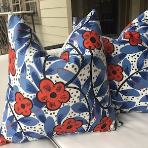 Könnte beinhalten: Zwei weiße Kissen mit einem blauen und roten Blumenmuster. Das Muster zeigt große rote Blumen mit schwarzen Zentren und blauen Blättern auf weißem Grund mit kleinen schwarzen Punkten.