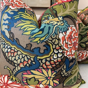 K&ouml;nnte beinhalten: Zwei dekorative Kissen mit braunem Hintergrund und einem farbenfrohen Blumen- und Drachenmuster. Die Kissen zeigen eine Mischung aus Rot, Orange, Gelb, Blau, Gr&uuml;n und Schwarz.