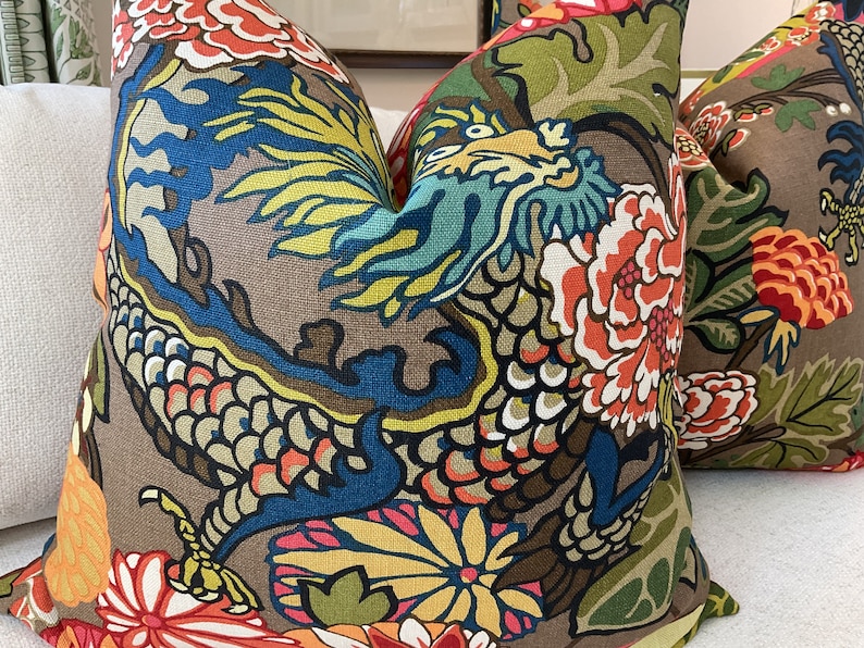 K&ouml;nnte beinhalten: Zwei dekorative Kissen mit braunem Hintergrund und einem farbenfrohen Blumen- und Drachenmuster. Die Kissen zeigen eine Mischung aus Rot, Orange, Gelb, Blau, Gr&uuml;n und Schwarz.